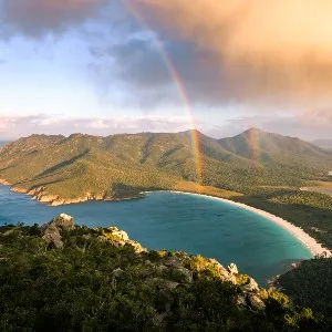 View of Wineglass Bay in Freycinet National Park. A storm cloud is passing over and a rainbow is shining down the middle of the picture leading into the water.