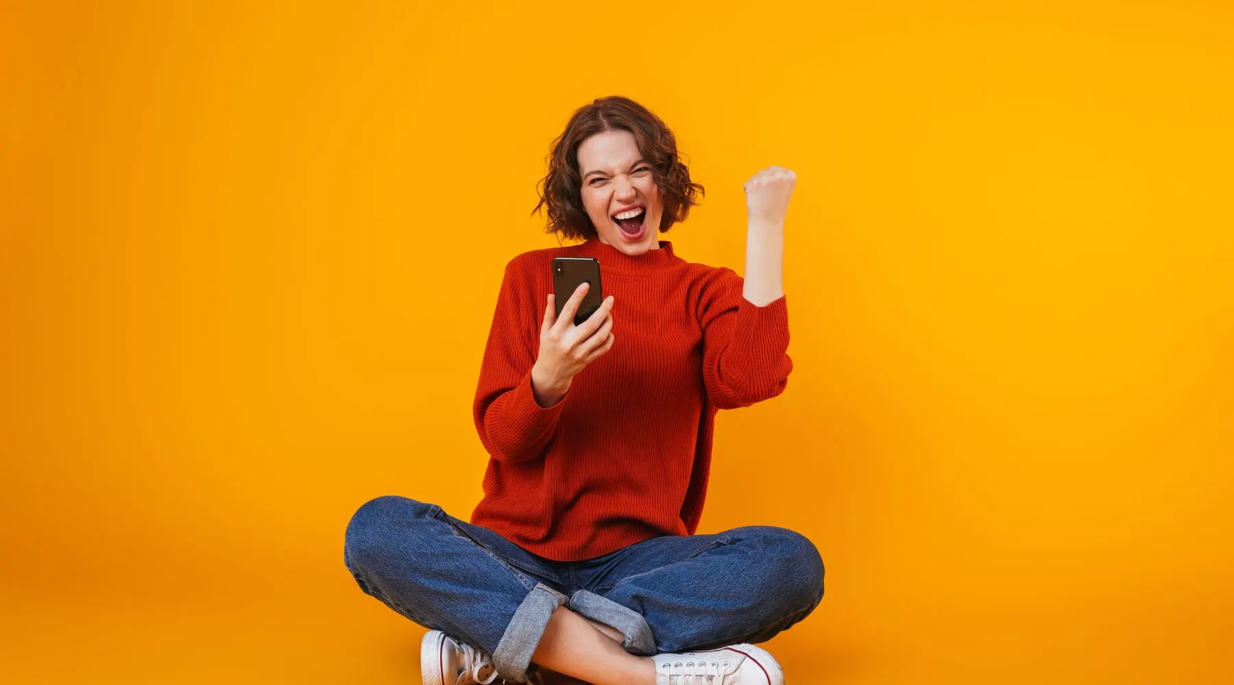 Image of young woman posing isolated over yellow wall background using mobile phone and making a winner gesture. 