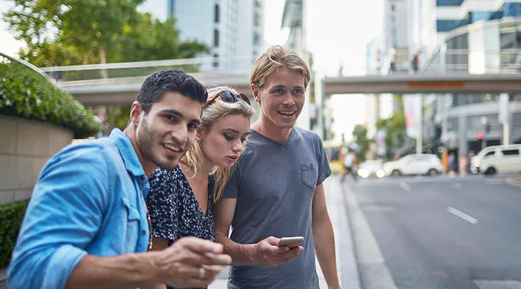 Three young friends waiting for a crowdsourced taxi in Sydney. Male is holding mobile phone and they are looking at street for the taxi.