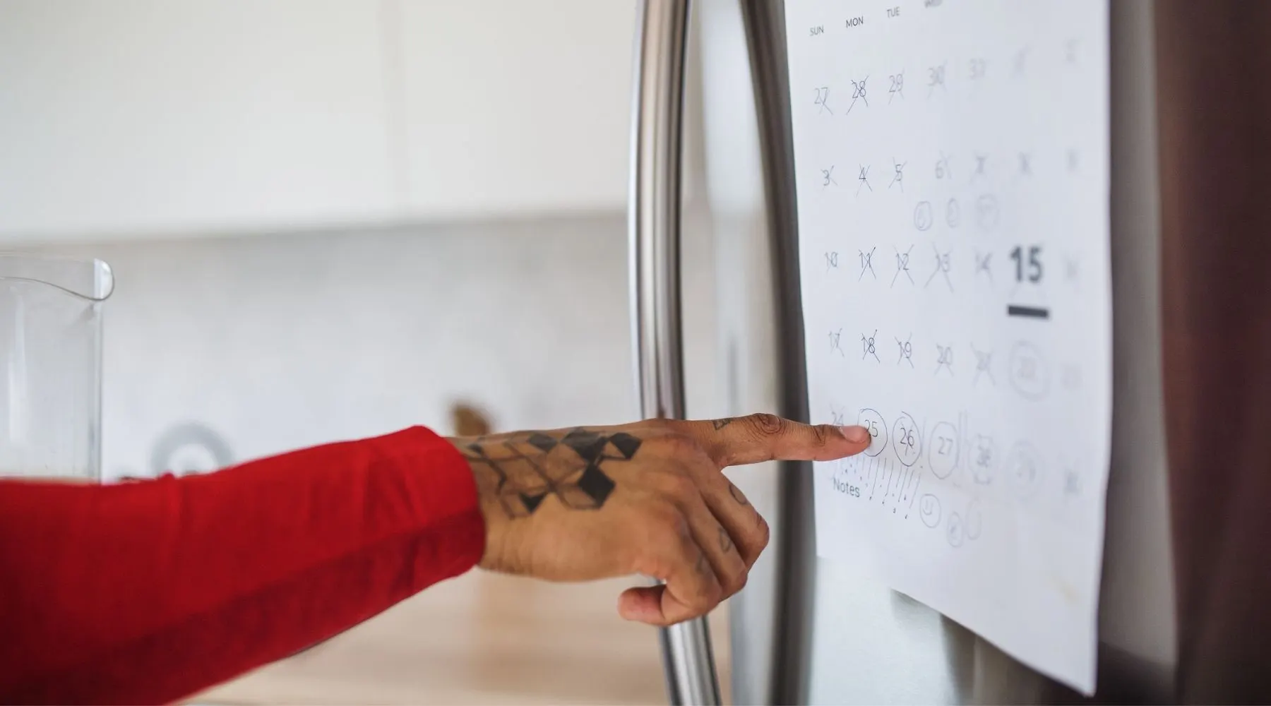 A man with a tattooed hand points at a calendar on the fridge.