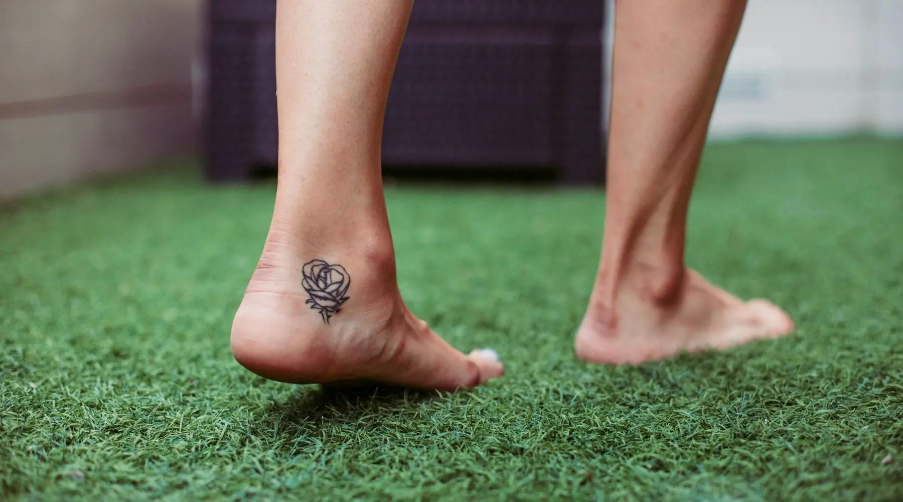 A woman with a tattoo of her rose on her foot walks on artificial grass.