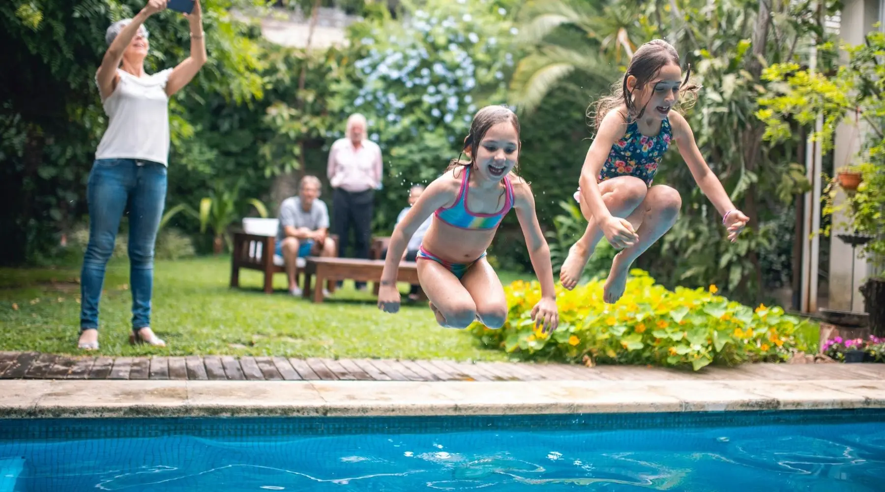 Two young girls jump into a family pool as an older relative takes photographs.