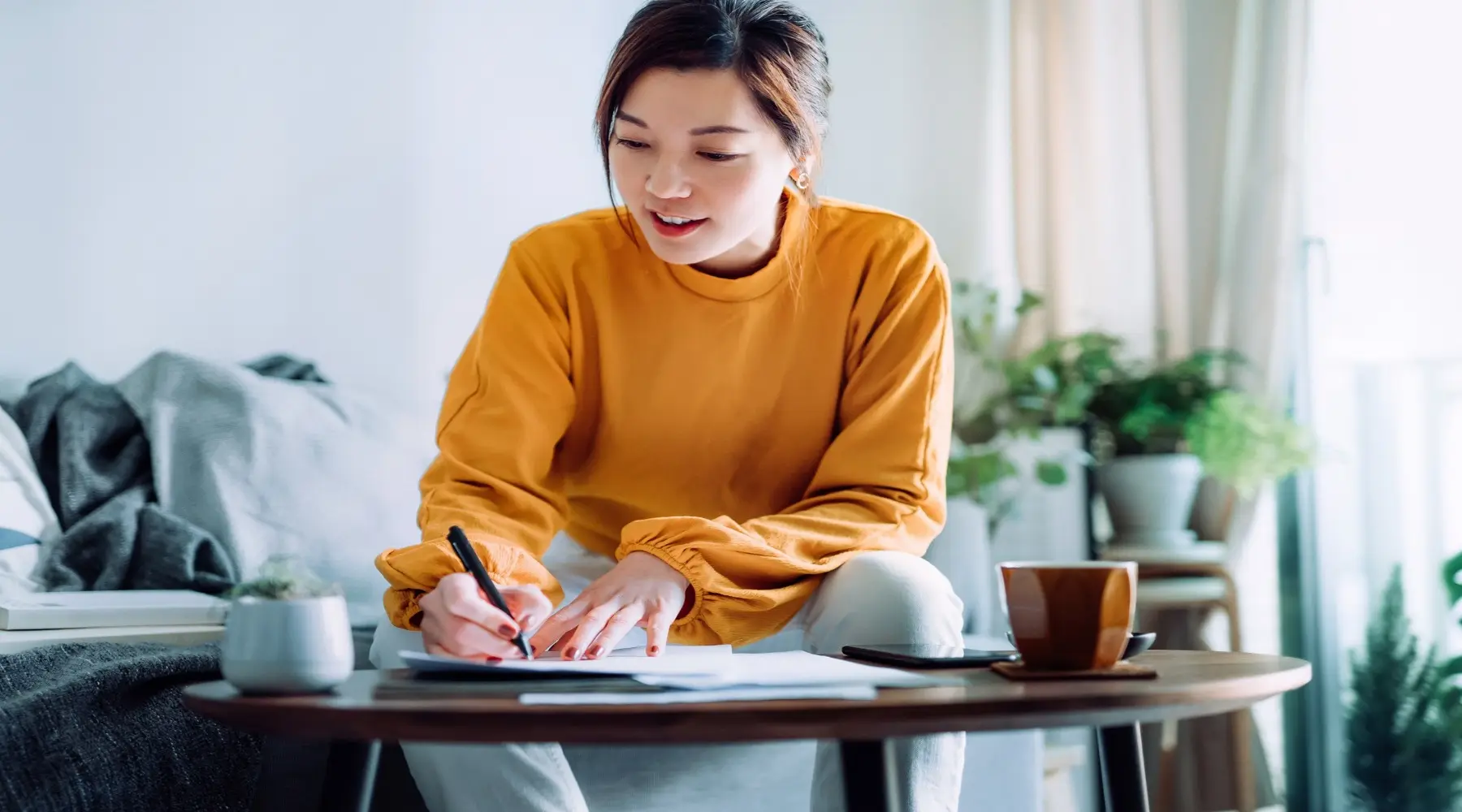 Young Asian woman holding a pen and signing paperwork in the living room at home_GettyImages_1800x1000