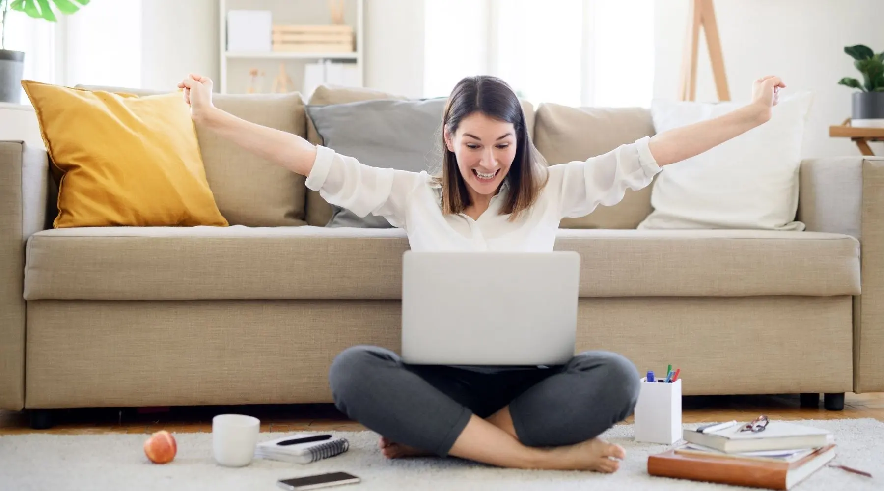 A young woman celebrates while sitting on the floor with her laptop.