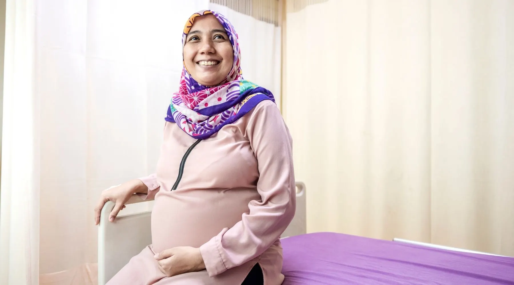 A pregnant lady wearing a religious headscarf sits on a hospital bed.