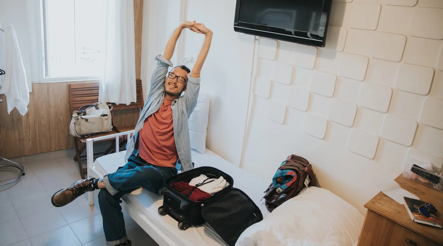 A smiling traveller stretching on a bed with a suitcase nearby.